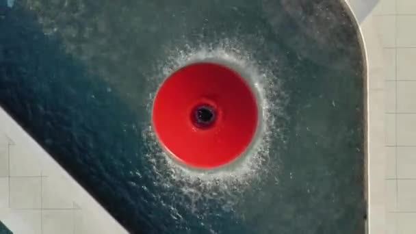 De drone stijgt langzaam uit boven een ronde rode fontein met spetterend water. Tafels staan in een halve cirkel rond de fontein — Stockvideo