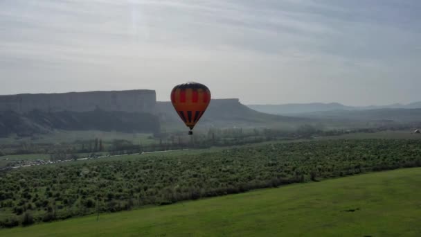 A multicolored hot air balloon with a basket flies against the blue sky. aerial view. — Stock Video