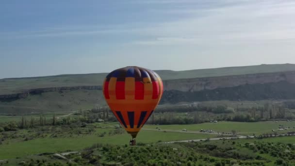 En flerfärgad varmluftsballong med en korg flyger mot den blå himlen. Flygbild. — Stockvideo