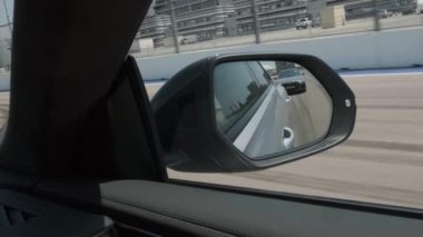 Hareket halindeki siyah araba, yan aynaya yansıyan yol, otomobilin uzun mesafeli aynası. Hareket halindeki arabanın arkasında başka bir araba daha var. Dikiz aynasından.