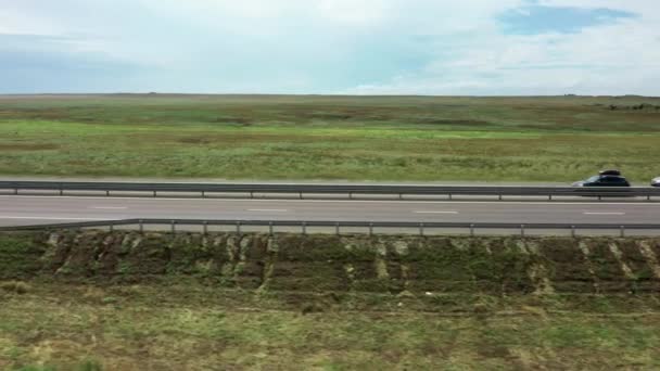 Los coches están conduciendo en una moderna carretera de varios carriles contra el fondo de un campo verde. Movimiento paralelo de la cámara. Grabación de video aéreo. Un día soleado. Transportes. — Vídeo de stock