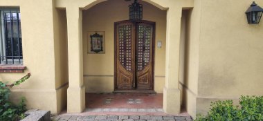 Sarı kır evinde dikdörtgen bir tasarım ve düz bir çatı var. Dış tarafında ahşap mobilyalar ve beyaz perdeli bir avlu bulunuyor. Şeffaf plastik örtüler. Çimenli ve ağaçlı yeşil arazi, sakin bir kırsal ortamı yansıtıyor.