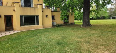 Sarı kır evinde dikdörtgen bir tasarım ve düz bir çatı var. Dış tarafında ahşap mobilyalar ve beyaz perdeli bir avlu bulunuyor. Şeffaf plastik örtüler. Çimenli ve ağaçlı yeşil arazi, sakin bir kırsal ortamı yansıtıyor.