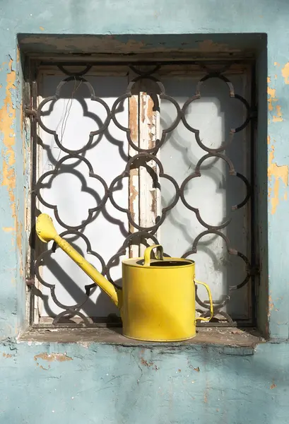 Lata de rega amarela na janela ralada da casa velha — Fotografia de Stock