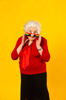 Sarı arka planda kırmızı gömlek ve gökkuşağı güneş gözlüğü takan yaşlı bir kadının stüdyo portresi.