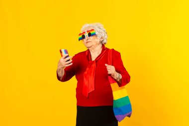 Kırmızı gömlek, gökkuşağı güneş gözlüğü ve çanta takan, cep telefonunu sarı arka planda gezdiren son sınıf bir kadının stüdyo portresi.