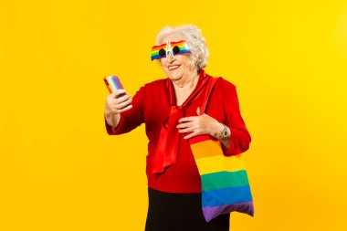 Kırmızı tişörtlü, gökkuşağı güneş gözlüklü ve çantalı, sarı arka planda cep telefonuyla video görüşmesi yapan son sınıf bir kadının stüdyo portresi.