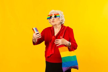 Kırmızı tişörtlü, gökkuşağı güneş gözlüklü ve çantalı, sarı arka planda cep telefonuyla video görüşmesi yapan son sınıf bir kadının stüdyo portresi.