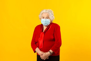 Kırmızı tişört giyen, sarı arka planda cerrahi maske takan yaşlı bir kadının stüdyo portresi.