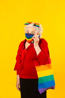 Kırmızı gömlek giyen, gökkuşağı güneş gözlüğü takan, gökkuşağı renkli yüz maskesi takan ve sarı arka planda cep telefonuyla konuşan son sınıf bir kadının stüdyo resmi.