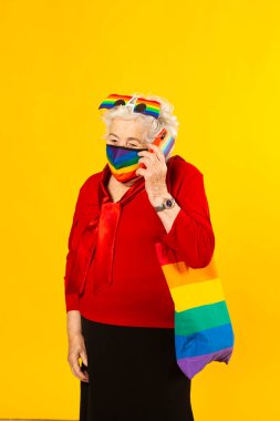 Kırmızı gömlek giyen, gökkuşağı güneş gözlüğü takan, gökkuşağı renkli yüz maskesi takan ve sarı arka planda cep telefonuyla konuşan son sınıf bir kadının stüdyo resmi.