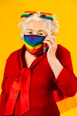 Kırmızı gömlek giyen, gökkuşağı güneş gözlüğü takan, gökkuşağı renkli yüz maskesi takan ve sarı arka planda cep telefonuyla konuşan son sınıf bir kadının stüdyo resmi.