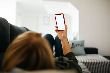 Orta yaşlı, gülümseyen sarışın bir kadının elinde bir kanepede yatarken elinde kırmızı bir akıllı telefon tutması.