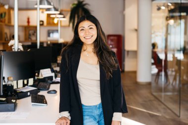 Kendine güvenen genç bir kadın kameraya gülümsüyor, aydınlık, modern bir ofiste duruyor.