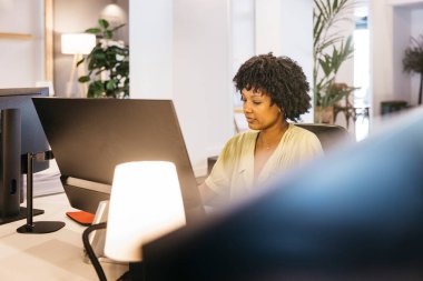Çağdaş bir ofis ortamında bilgisayar masasında işine konsantre olan siyah bir kadın.