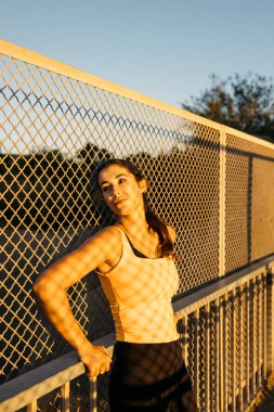 Kadın sporcu gün batımında çitlere karşı egzersiz yaptıktan sonra duraklıyor