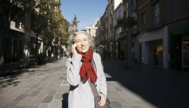 Şehir caddesinde yürüyen son sınıf öğrencisi bir kadın, cep telefonuyla konuşurken gülümsüyor.