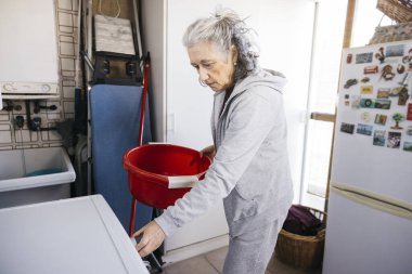 Yaşlı kadın çamaşır yıkıyor, çamaşır makinesine kırmızı leğen koyuyor.