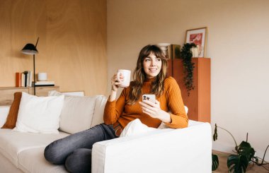 Kanepede dinlenen, akıllı telefon kullanan ve oturma odasında kahve içen bir kadın.