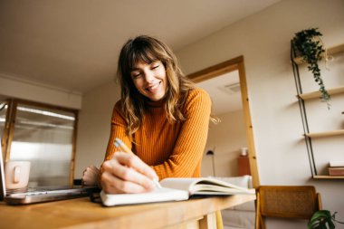 Gülümseyen bir kadın, evdeki ofis masasında dizüstü bilgisayarla bir deftere yazı yazıyor.