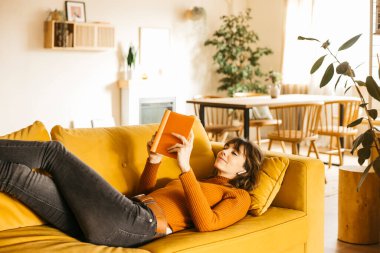 Sarı bir kanepede uzanmış, rahat oturma odasında kitap okuyan bir kadın.