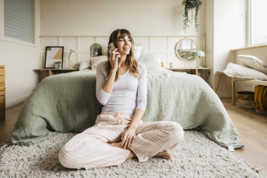 Modern yatak odasında yerde oturan pijamalı bir kadın cep telefonuyla konuşuyor.