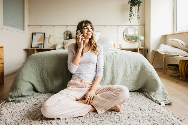 Modern yatak odasında yerde oturan pijamalı bir kadın cep telefonuyla konuşuyor.