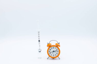 Şırıngalar, Covid-19 'a karşı bir aşı şişesine yerleştirilmiş. Yanında da zaman kavramı olarak alarm saati var. Aşılanması gereken ve ikinci dozun alınması için, izole edilmiş..