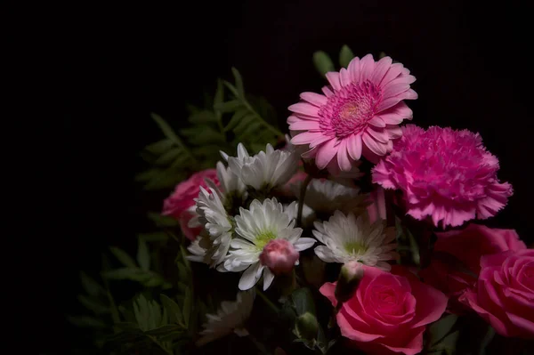 Bir buket pembe karanfil ve beyaz kasımpatı ve siyah arka planda pembe gerberalar olan bir gül.