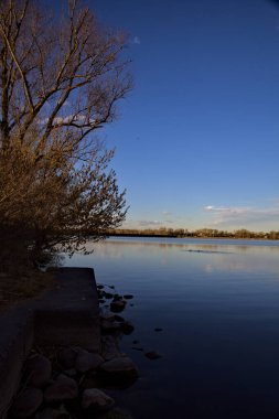 Gün batımında bir gölün kıyısında, arkasında ağaçlar ve dağlar var.
