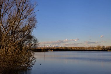Gün batımında bir gölün kıyısında, arkasında ağaçlar ve dağlar var.