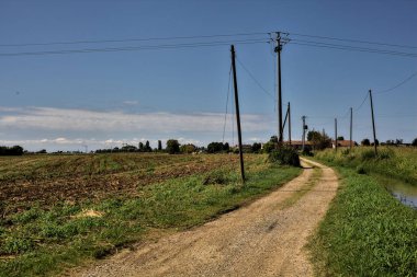 Açık bir günde İtalya kırsalında tarlaların yanında toprak yol ve bir su deresi.
