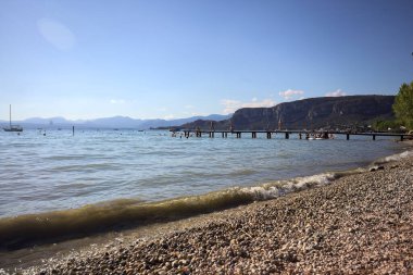 Güneşli bir günde insanların oturduğu bir gölün üzerinde uzanan iskele ve arka planda bir dağ sırtı.