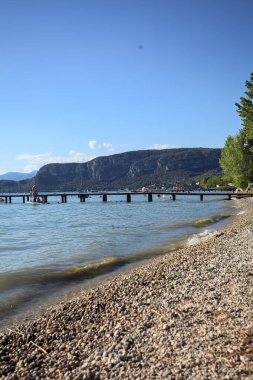 Güneşli bir günde insanların oturduğu bir gölün üzerinde uzanan iskele ve arka planda bir dağ sırtı.