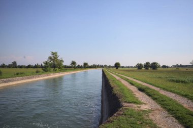 İtalya 'nın kırsal kesimlerinde güneşli bir günde çayırın ortasında patikalarla çevrili bir kanalda akan sular.