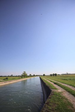 İtalya 'nın kırsal kesimlerinde güneşli bir günde çayırın ortasında patikalarla çevrili bir kanalda akan sular.