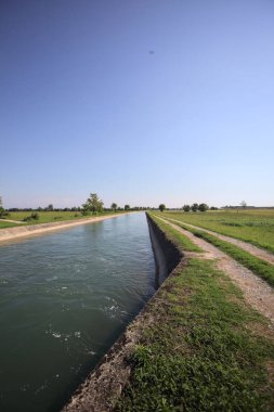 İtalya 'nın kırsal kesimlerinde güneşli bir günde çayırın ortasında patikalarla çevrili bir kanalda akan sular.
