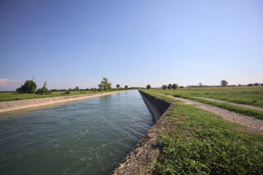 İtalya 'nın kırsal kesimlerinde güneşli bir günde çayırın ortasında patikalarla çevrili bir kanalda akan sular.