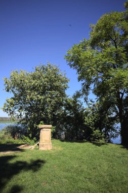 Stone pillar and trees on a lawn next to the lakeshore on a sunny day