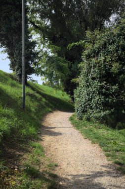 Narrow passage on a trail passing between an embankment and a stone rail with a hedge behind it in a park