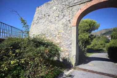 Remains of a stone wall with a gate on a trail between hedges in a park on a sunny day with a clear sky as background