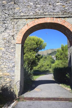 Remains of a stone wall with a gate on a trail between hedges in a park on a sunny day with a clear sky as background