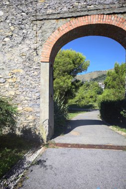 Remains of a stone wall with a gate on a trail between hedges in a park on a sunny day with a clear sky as background