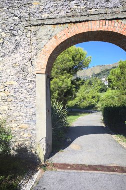 Remains of a stone wall with a gate on a trail between hedges in a park on a sunny day with a clear sky as background