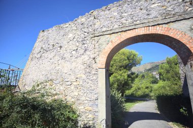 Remains of a stone wall with a gate on a trail between hedges in a park on a sunny day with a clear sky as background