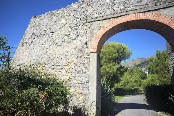 Remains of a stone wall with a gate on a trail between hedges in a park on a sunny day with a clear sky as background