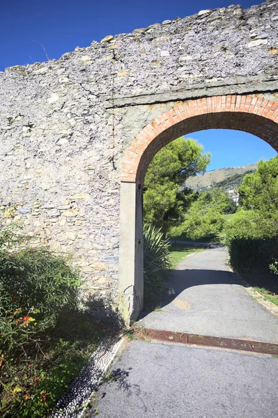 Remains of a stone wall with a gate on a trail between hedges in a park on a sunny day with a clear sky as background