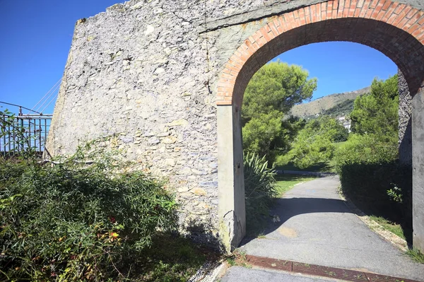 Remains of a stone wall with a gate on a trail between hedges in a park on a sunny day with a clear sky as background