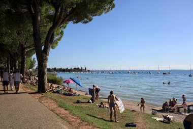 Malcesine, İtalya - 31. AĞUSTOS 2025 - İnsanlar, Garda Gölü kıyısındaki ağaçların olduğu bir sahilde güneşlenip, yatıyorlar.