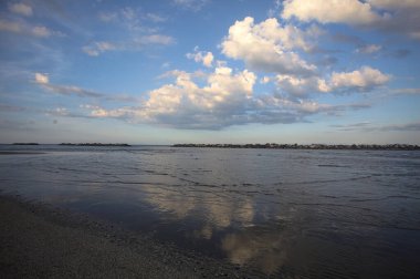 Gün batımında sahilde dalgalı bir su ve kısmen bulutlu bir gökyüzü suya yansıyor.